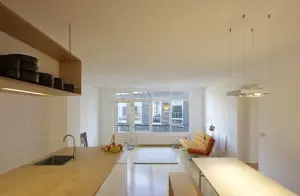 8.Dooghe: ‘Multiplex verbindt alles in huis. Het ligt op de vloer en ook keuken, banken en zijkanten van kasten en nissen zijn ervan gemaakt. Het is een mooi en makkelijk te verwerken materiaal, dat met een laag vernis goed tegen water kan.’ Het keukenblok is efficiënt ingericht met greeploze Ikea kastjes aan de eetkamerkant en koelkast, afwasmachine en schuifkasten aan de kookkant. De hangende bovenkast is voor de ene helft open servieskast en voor de andere helft voorraadkast en behuizing van de afzuigkap. De inrichting is rustig en open gehouden. De rechthoekige lamp boven de eettafel is een Grand Prix van Delta Light. In de woonkamer staan een Artifort bank van Kho Liang Ie en een schommelstoel van Charles Eames. De vintage vloerlamp vond Dooghe in een vorige woning en de lamp boven het vloerkleed is een ontwerp van Philippe Starck.