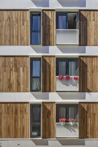 Uitkragende kaders van wit aluminium rondom de ramen creëren dieptewerking met schaduwen en reliëf op de vlakke gevel. Kijk je recht op de gevel, dan zie je vooral de houten bekleding. Kijk je er zijdelings op, dan is alles wit. Daarnaast zorgen de kaders van binnenuit voor extra stevigheid en geborgenheid