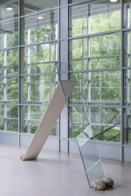 Het werk Standing (1969/2025) staat voor een reeks plafondhoge ramen. Een glazen plaat leunt tegen een houten paneel, en vier stenen houden het geheel in balans. 
