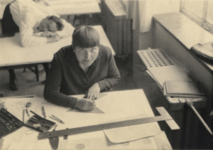 Lotte Stam Beese aan de tekentafel met Helmut Schulze op de achtergrond. 